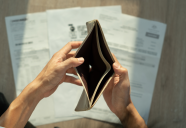 Hands opening an empty wallet. Paper bills pilled up on the table in the background.